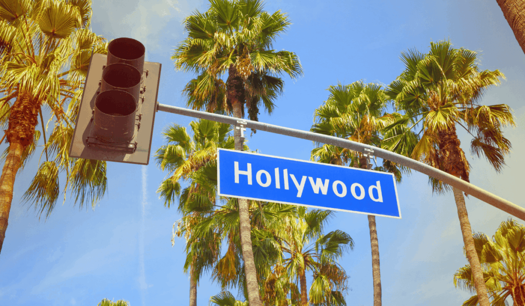 Hollywood feliratú útjelző tábla pálmafák és közlekedési lámpa között, napsütéses kaliforniai hangulatban.