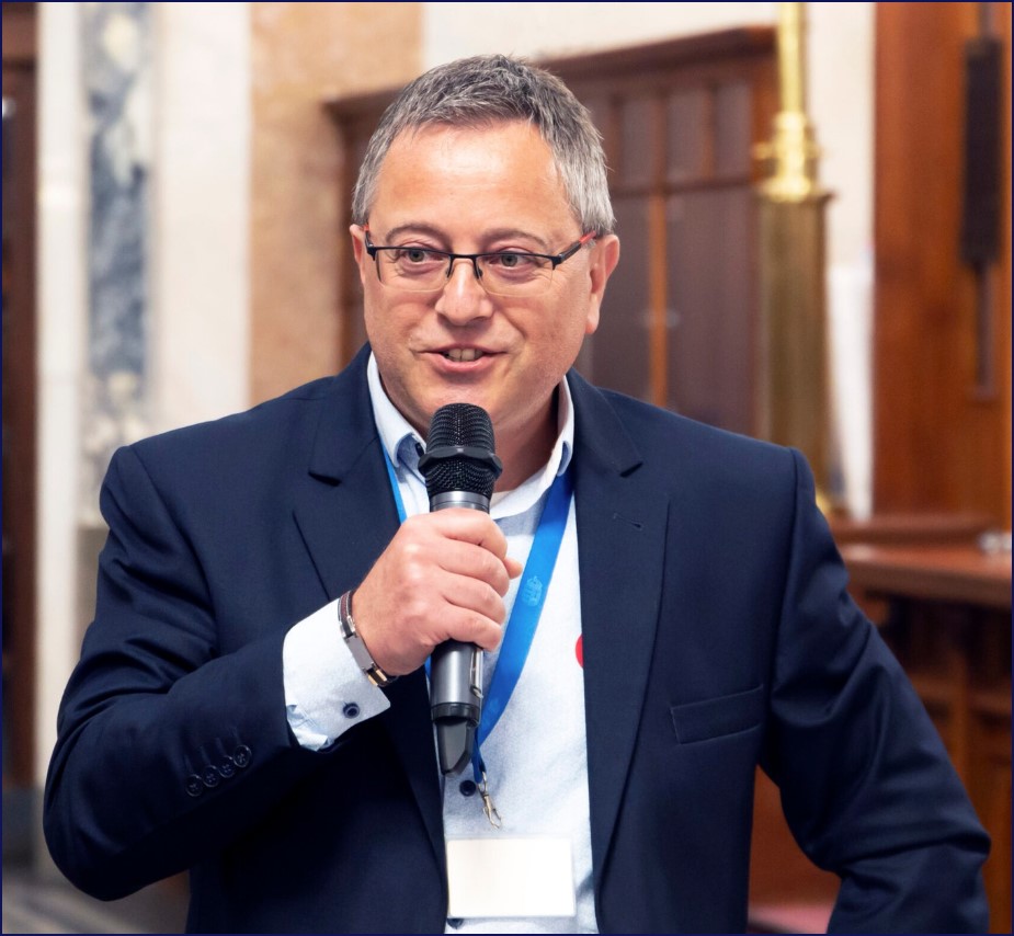 Tisótzki István mikrofont tartva beszél egy szakmai rendezvényen, sötét zakóban és kék nyakpánttal.