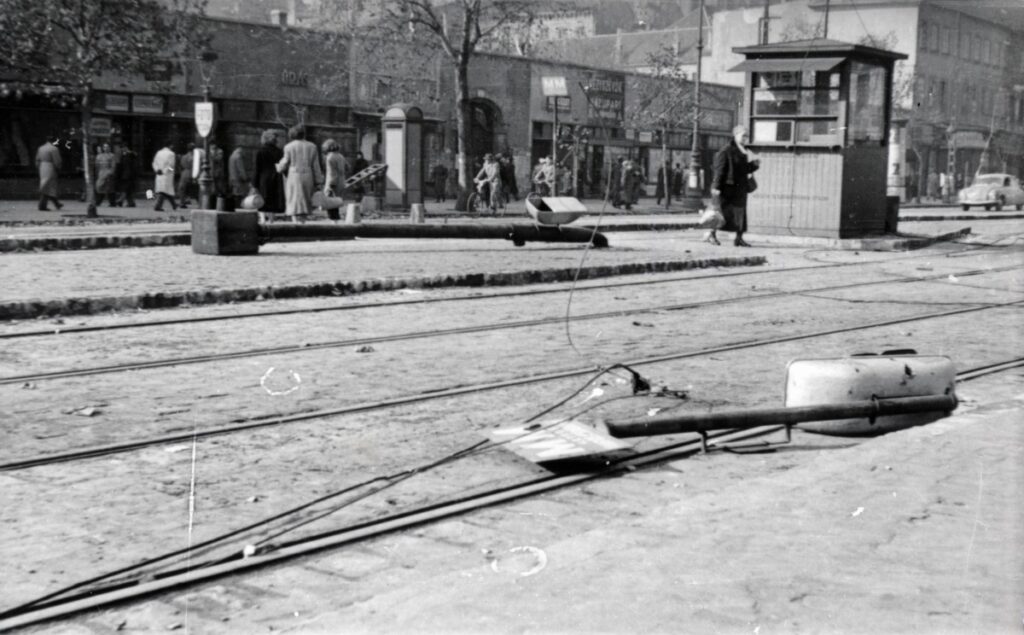 Kidöntött oszlopok és vezetékek a Károly körút villamossínjein, háttérben járókelőkkel, 1956-ban.