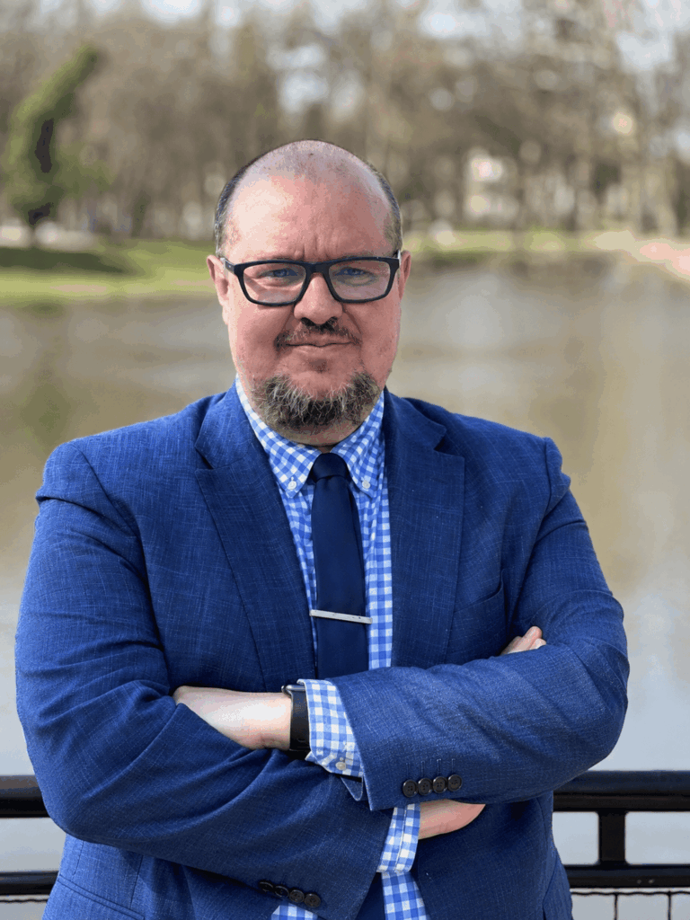 Bányász Péter kék zakóban, szemüvegben, összefont karral, háttérben fákkal és vízparttal.
